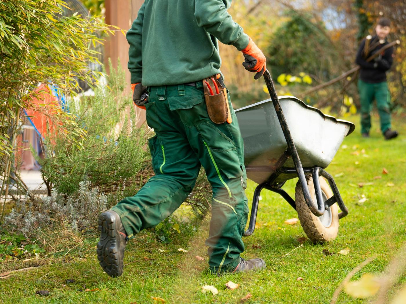 Two crew members in green work clothes moving a wheelbarrow across a lawn.